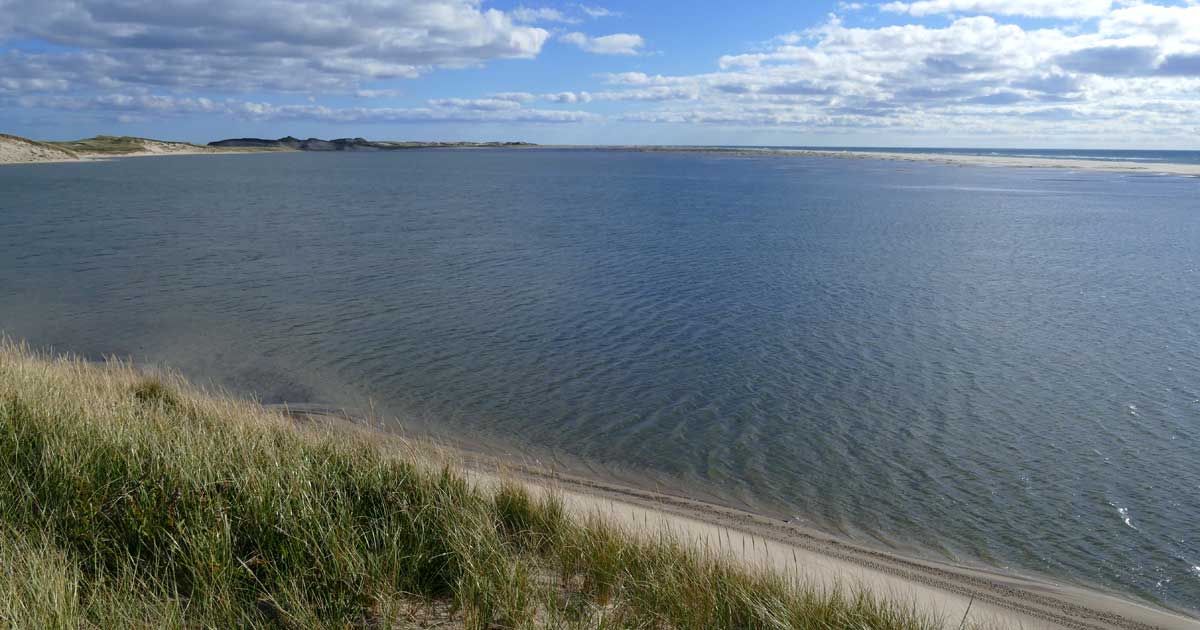 A vast expanse of flat sandy beach entirely underwater, with a line of high dunes along one side and a thin strip of sand along the opposite side. The strip is the only dry land between the floodwater and the ocean.