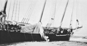 The hull of a grounded schooner lying at an angle on the beach, with some masts and rigging still intact, and a tumble of sail and rope hanging over the side.